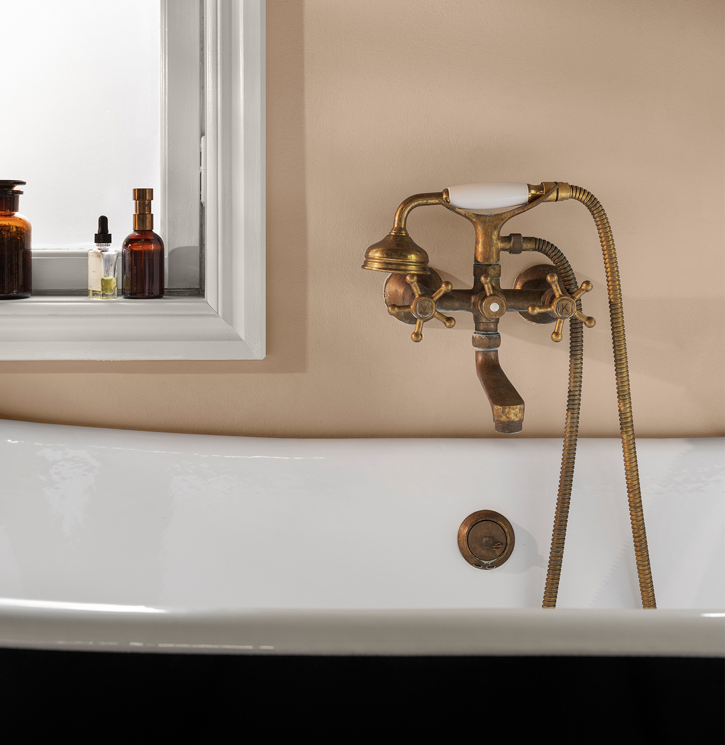 Vintage badekar med en antikk messingkran og dusjhode, mot en rosabeige vegg malt i LADY Aqua våtromsmaling i fargen Jotun Devine og et vindu med flasker på karmen