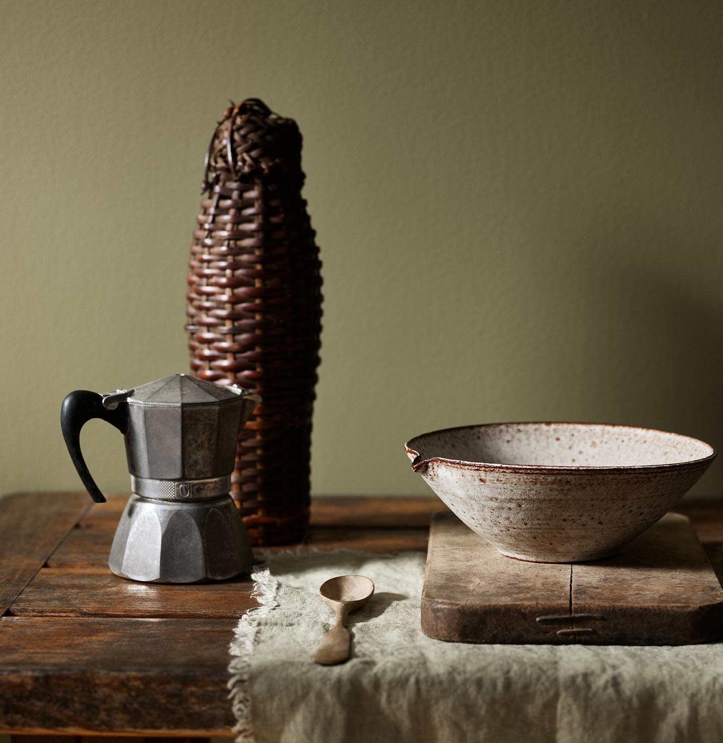 Rustic wooden table with a metal espresso maker and ceramic bowl with a olive green background painted with the Jotun colour 8597 Seaweed Green.