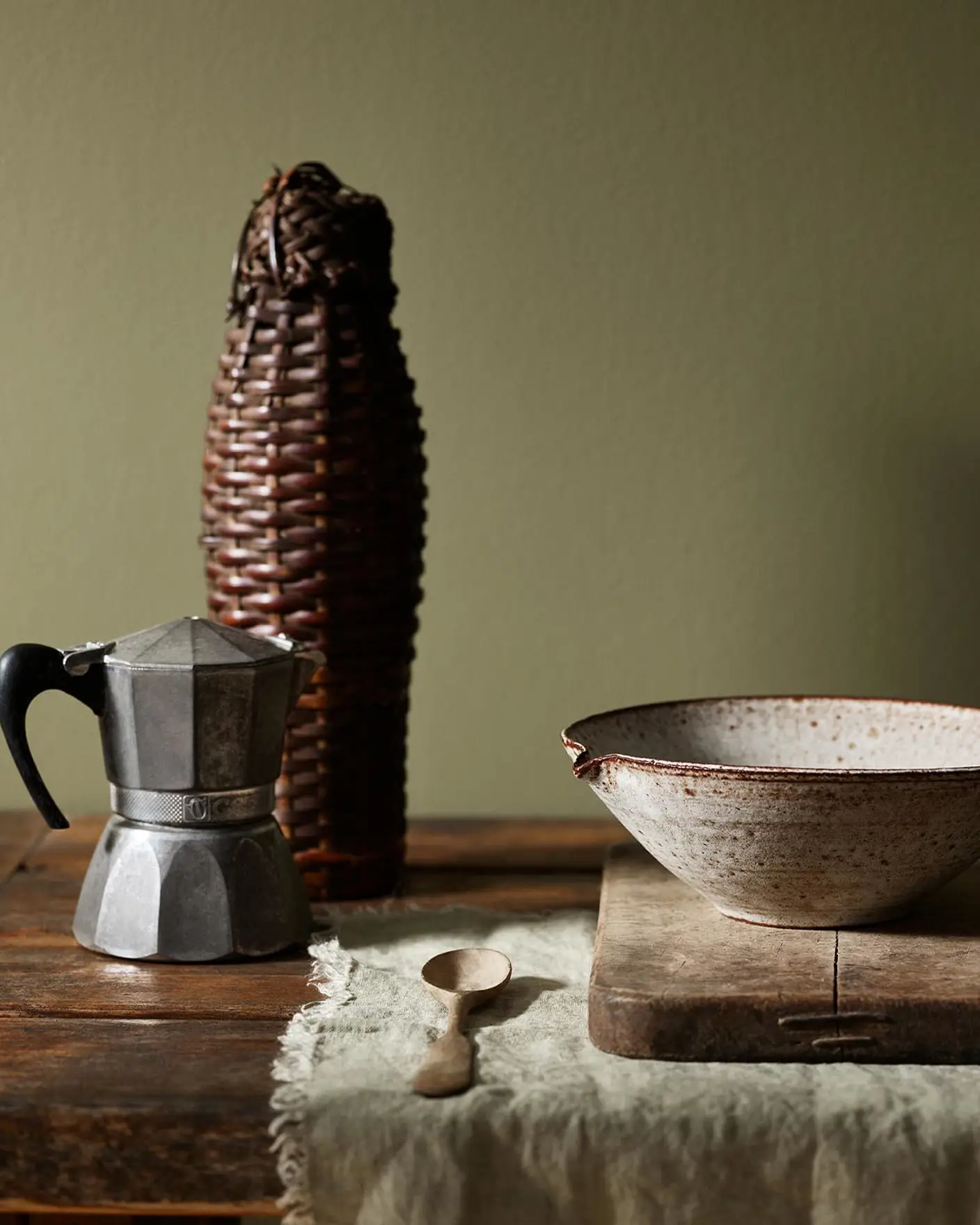 Rustic wooden table with a metal espresso maker and ceramic bowl with a olive green background painted with the Jotun colour 8597 Seaweed Green.