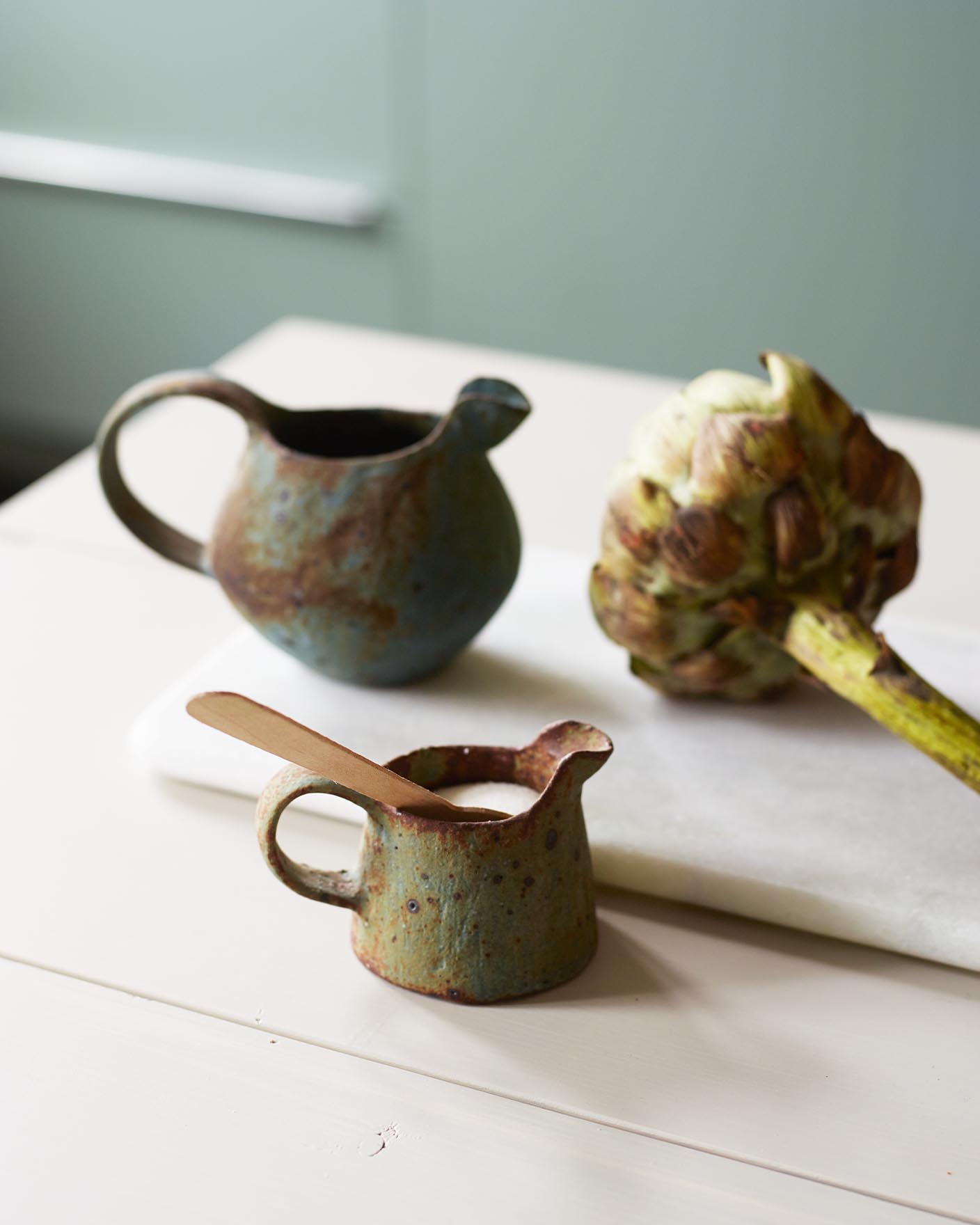 Rustikk keramikk og en frisk artisjokk hviler på en lys overflate, med myke grønne vegger malt med Jotun 7163 Minty Breeze som gir scenen en kjølig og forfriskende bakgrunn.