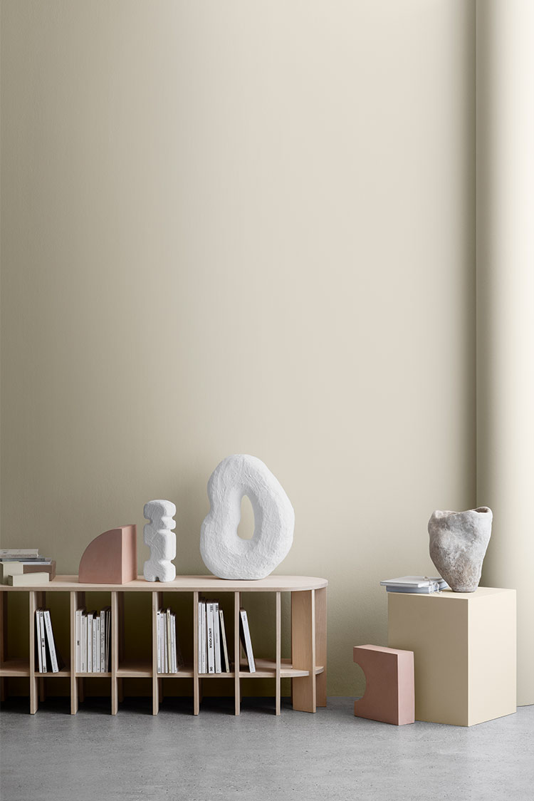 Moderne minimalistisk interiør med abstrakte skulpturer på beige hyller, bøker organisert vertikalt, og naturlig lys på en greige vegg malt i Jotun 12182 Gente Whisper