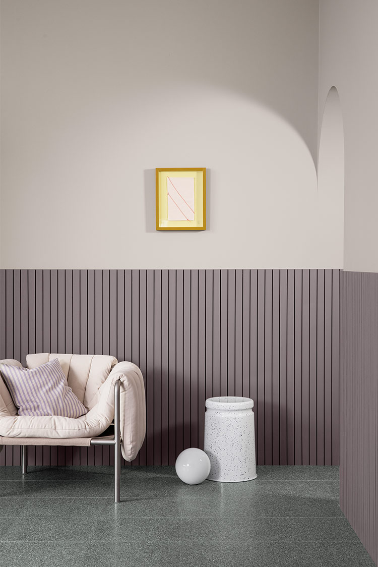 Moderne stueinteriør med en kremfarget lenestol, stripete pute, flekket vase og en hvit sfærisk dekorasjon mot en grålilla vegg malt i Jotun 3377 Slate Lavender med vertikale paneler og innrammet abstrakt kunst