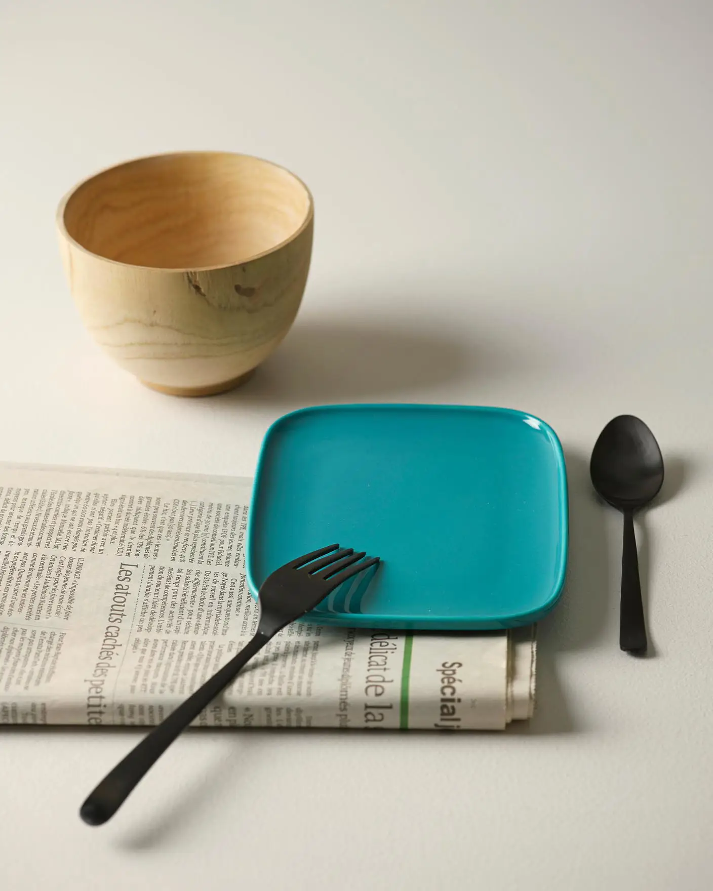 Una mesa con un plato color teal, cubiertos negros, un cuenco de madera y un periódico sobre un fondo blanco dorado claro pintado con Jotun 1624 Skylight.