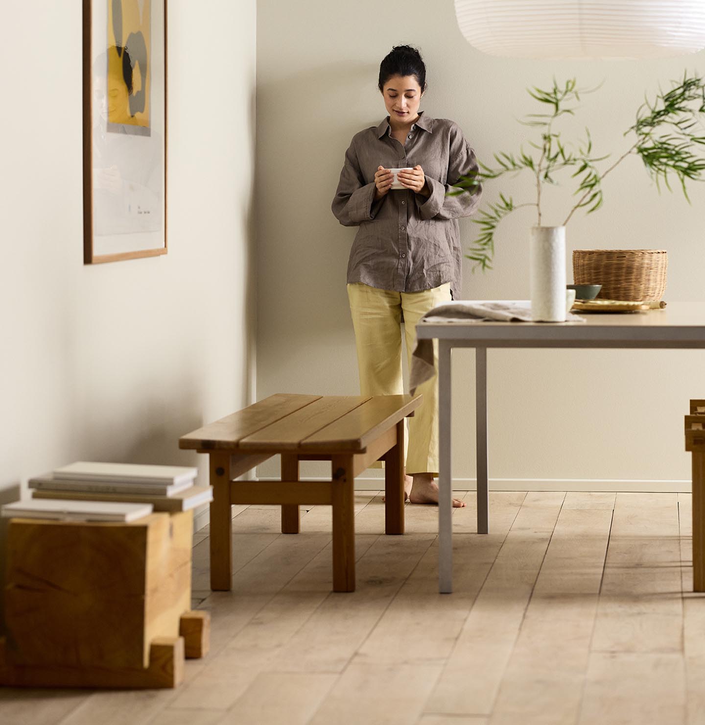 Woman in a minimalist room with wooden bench, table decor, and framed artwork against a beige wall painted in the Jotun colour 12308 Unbleached.