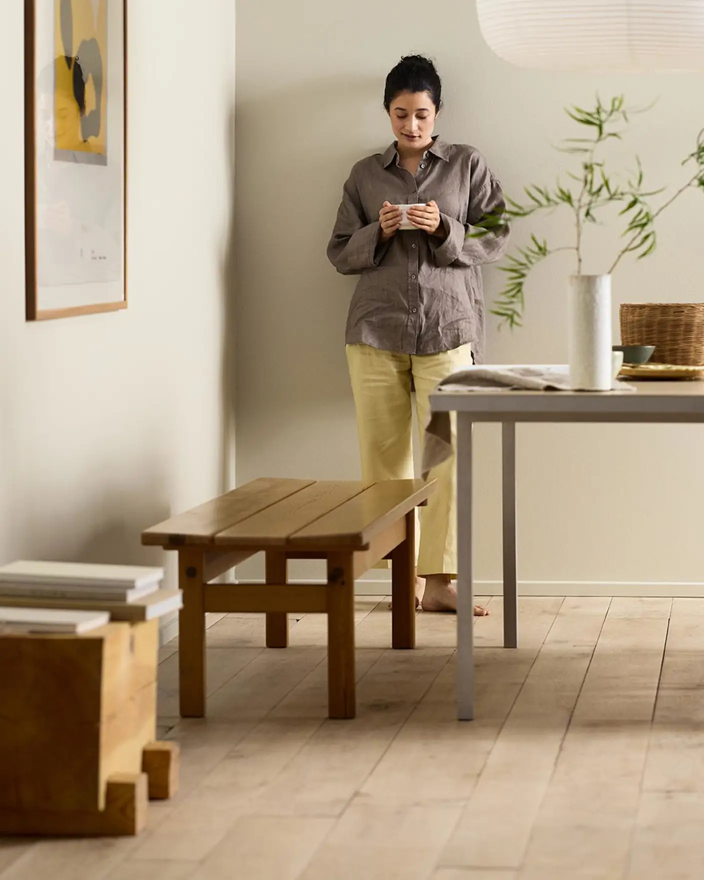 Woman in a minimalist room with wooden bench, table decor, and framed artwork against a beige wall painted in the Jotun colour 12308 Unbleached.