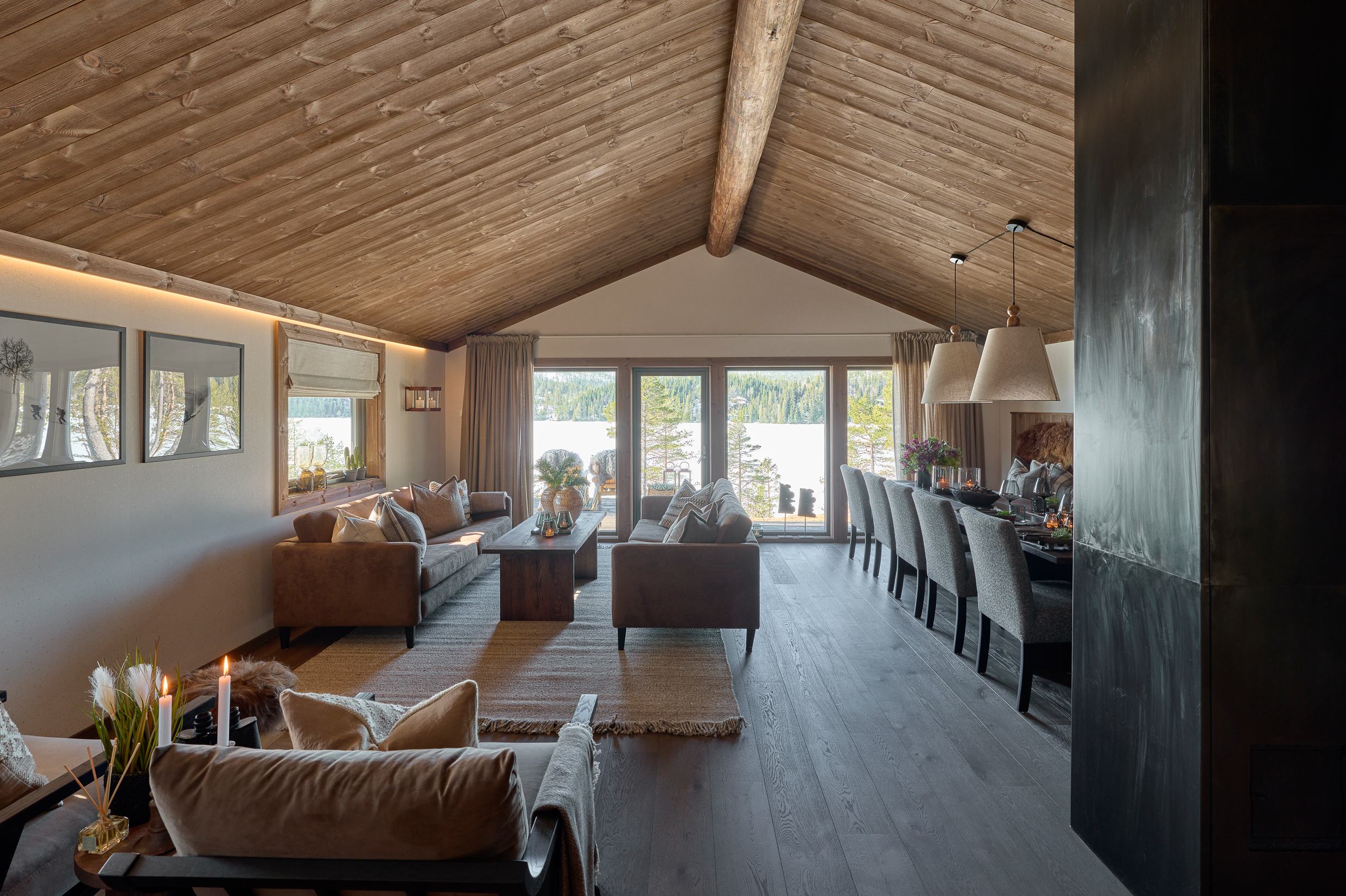 Stue sett mot utsikten, mørkt trepanel i tak, hvitt tapet, lun stemning med mørke møbler.