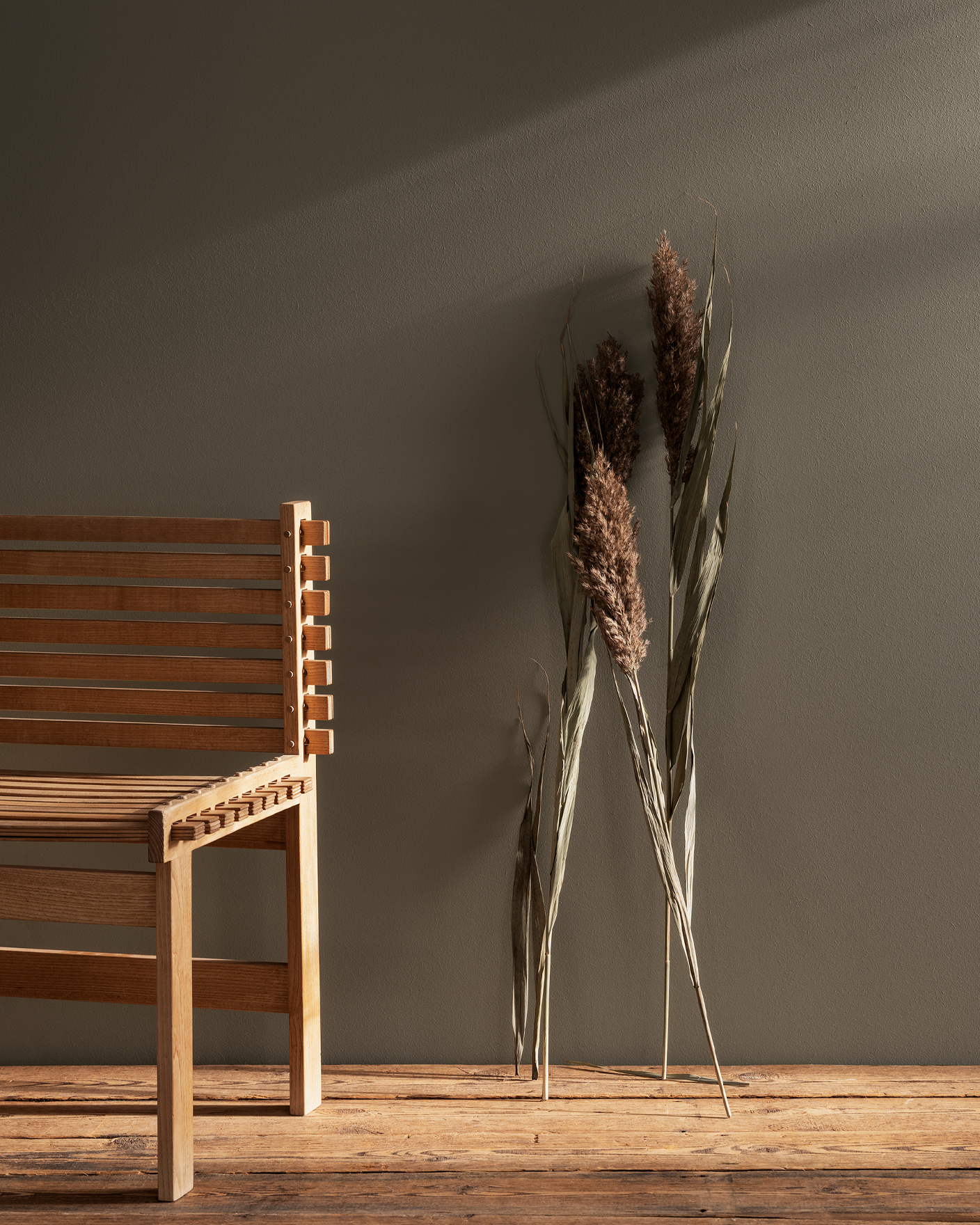 Träbänk i minimalistisk design bredvid torkat gräs mot en neutral vägg målad Jotun 11012 Rustic Brown. 