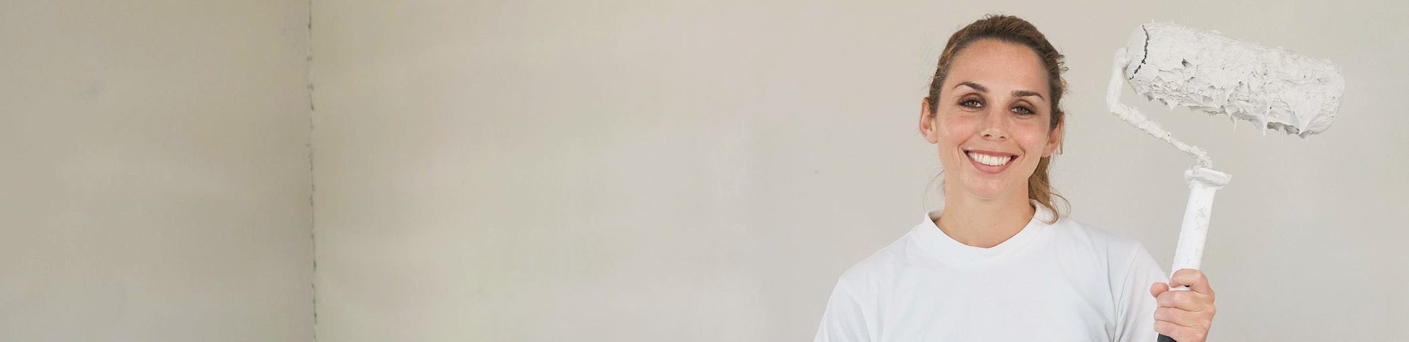 Kvinne som holder en malerulle mot en hvit vegg, smiler iført hvite klær, klar til å rullesparkle