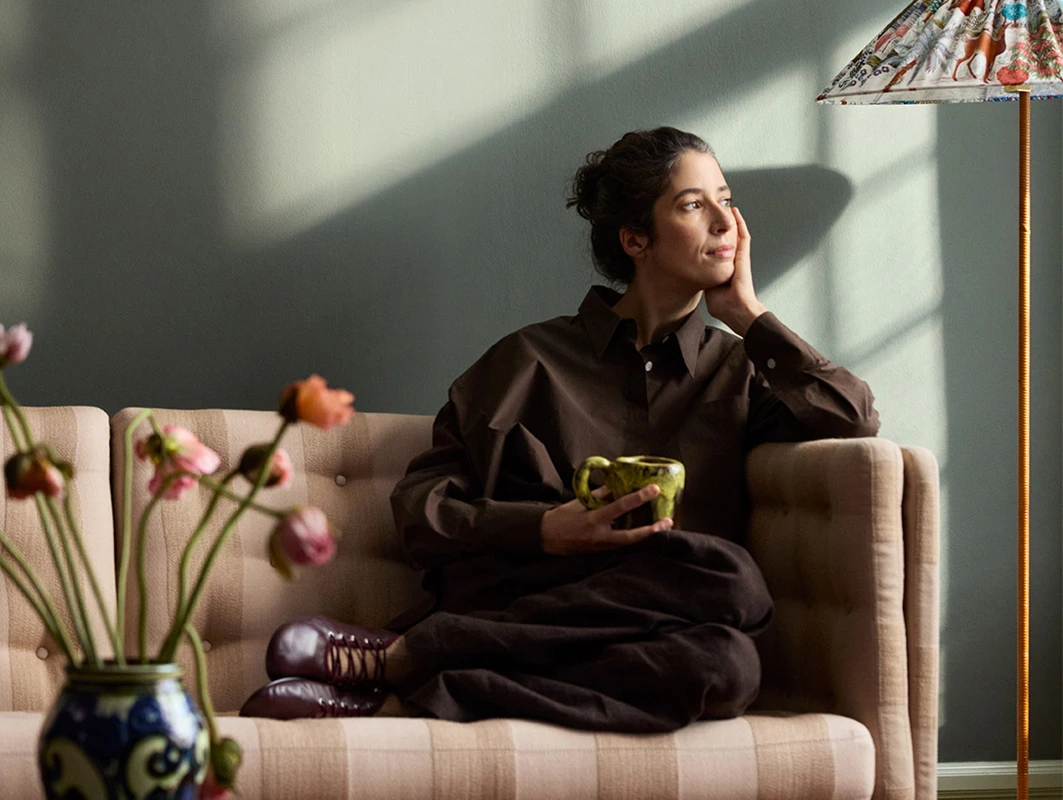 Woman sitting on a beige tufted sofa against a Jotun Northern Mystic wall, holding a green mug; floral lamp and vase add warmth.