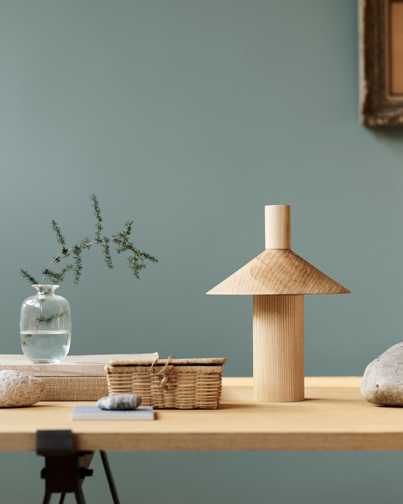 Minimalist decor with vase, books, and sculpture on a wooden table against a muted, bluish green background in the colour Jotun 6379 Cityscape.