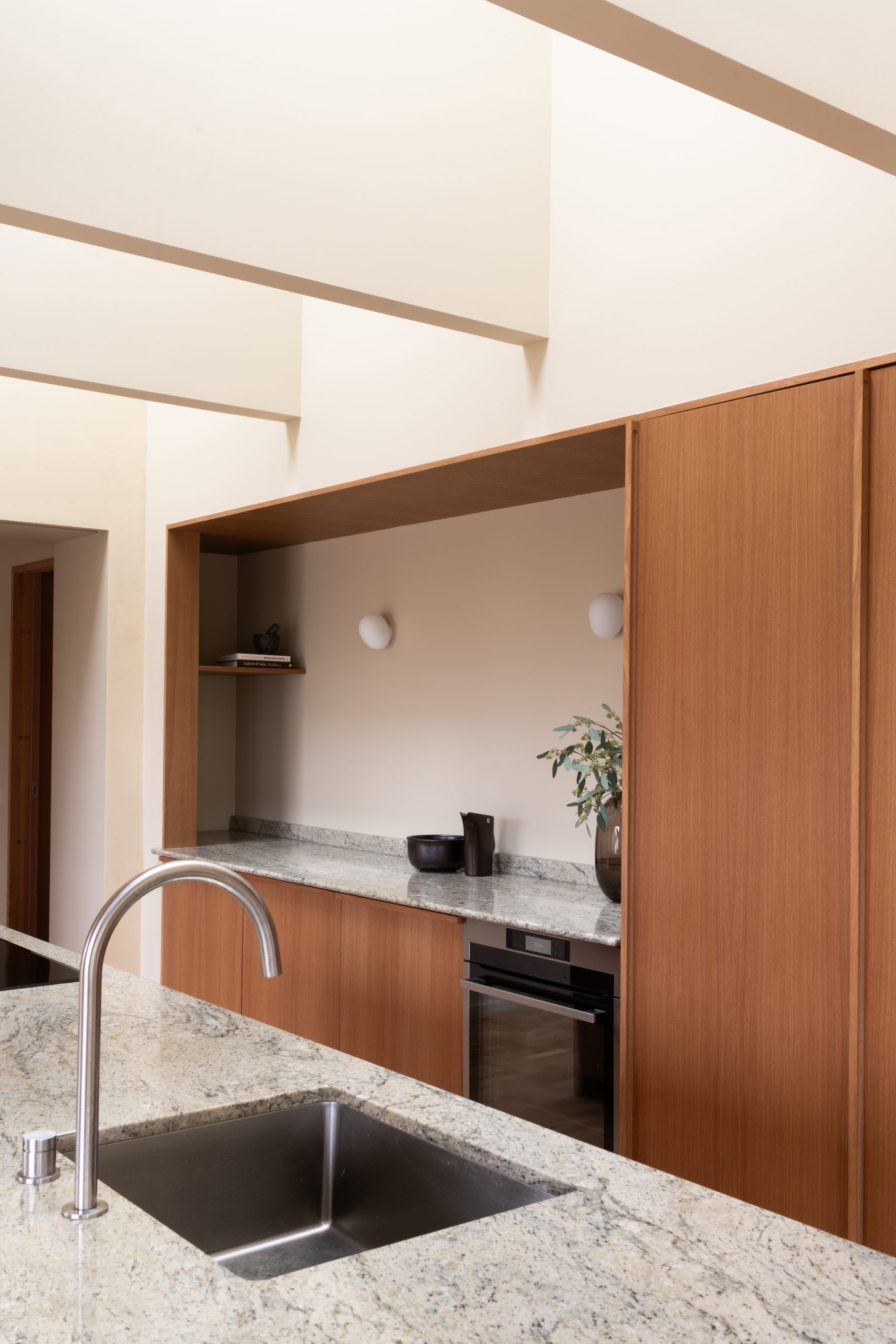 Moderne kjøkkeninteriør med slank, rustfri kjøkkenkran og vask i forgrunnen. Benkeplaten er laget av lys granitt, og skapene har et stilrent trefinish. En ovn er integrert i skapene, og en plante står på benken som dekor. Vegger og tak malt i gulbeige med Jotun 11173 Humble Yellow