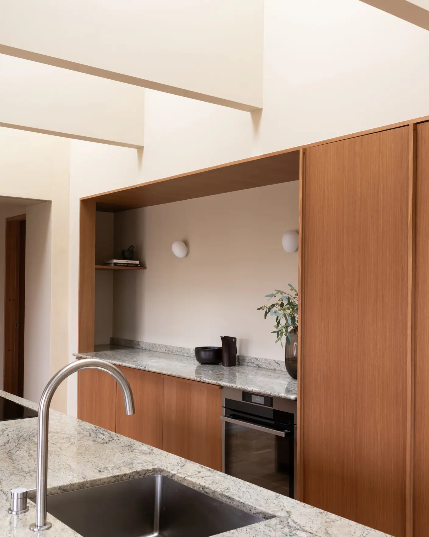 Moderne kjøkkeninteriør med slank, rustfri kjøkkenkran og vask i forgrunnen. Benkeplaten er laget av lys granitt, og skapene har et stilrent trefinish. En ovn er integrert i skapene, og en plante står på benken som dekor. Vegger og tak malt i gulbeige med Jotun 11173 Humble Yellow