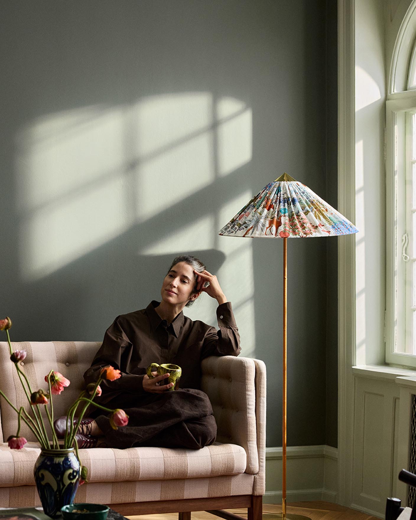 Woman sitting in a sofa with sunlight streaming through a window. The background wall is painted with the dark, muted green colour 7613 Northern Mystic from Jotun.