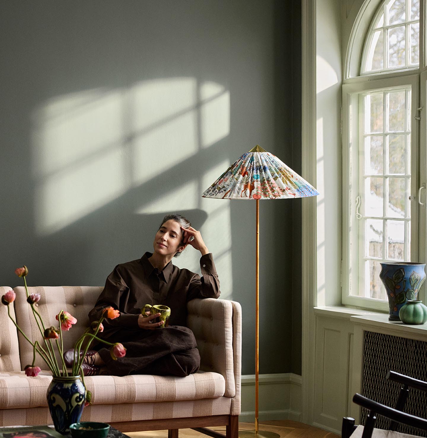 Woman sitting in a sofa with sunlight streaming through a window. The background wall is painted with the dark, muted green colour 7613 Northern Mystic from Jotun.