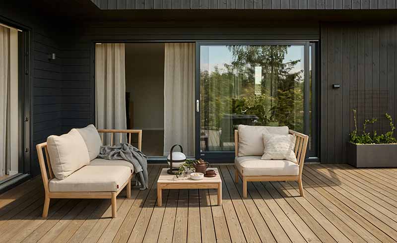 Moderne terrasse med lyst brune terrassebord i tre som er beiset med TREBITT Terrasse i fargen 9074 Nordisk Tre, to komfortable lenestoler med lys beige puter, et lite trebord med te-sett, og store glasskyvedører som fører inn til huset som er malt i Jotun 90033 Funkissort