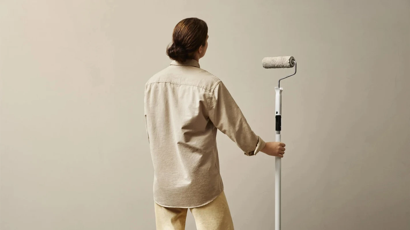 Kvinne står foran en vegg hun skal male og holder en malerulle med skaft