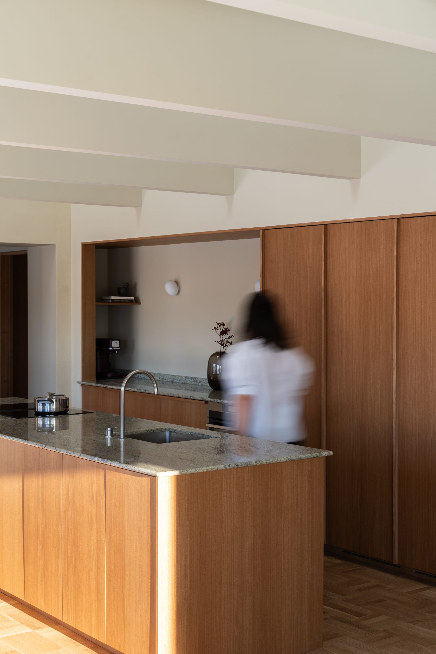 Moderne kjøkkeninnredning i tre med marmorbenk, integrerte skap, vegger og tak malt i Jotun 11173 Humble Yellow og minimalistisk design. Utydelig person beveger seg forbi kjøkkenøyen