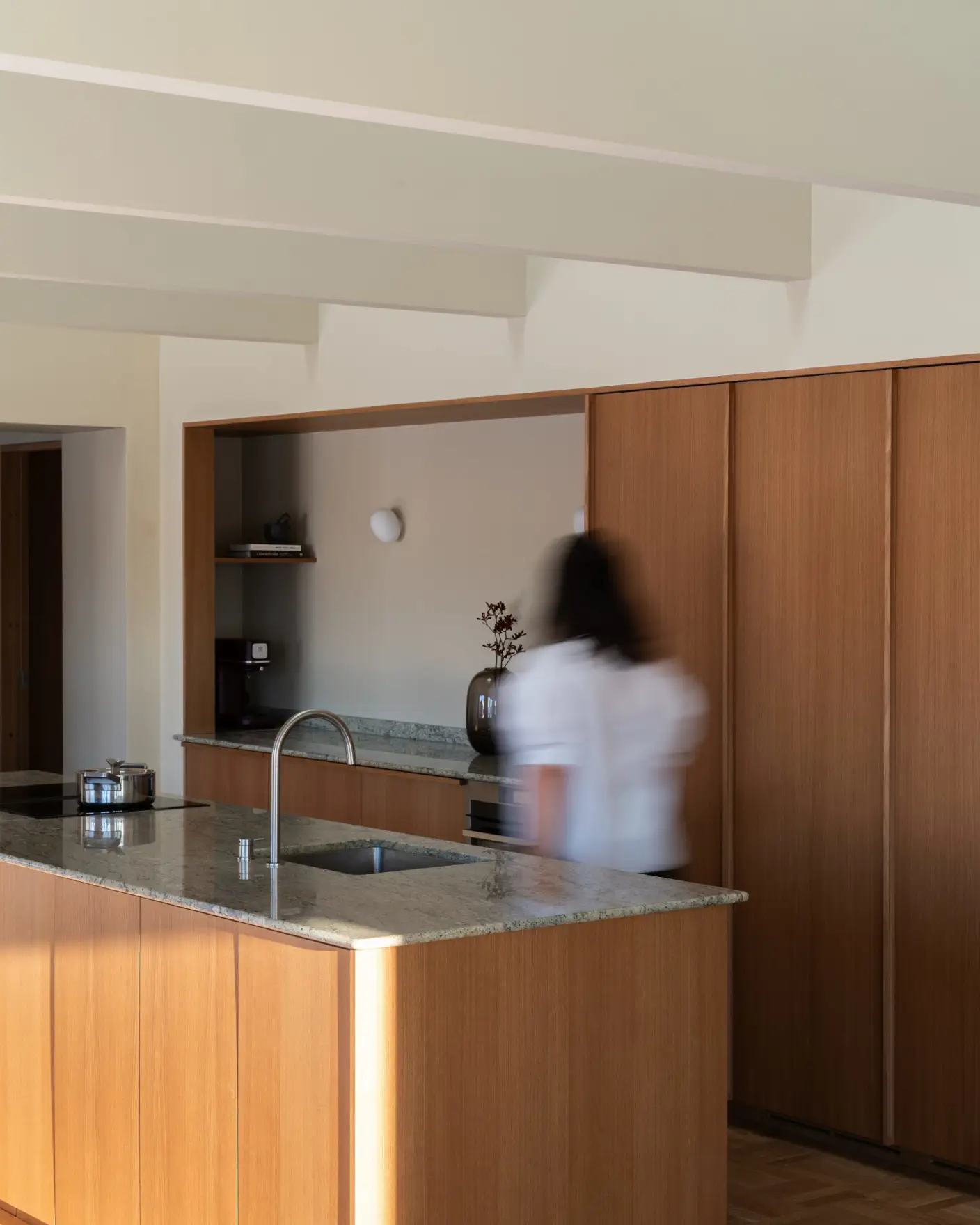 Moderne kjøkkeninnredning i tre med marmorbenk, integrerte skap, vegger og tak malt i Jotun 11173 Humble Yellow og minimalistisk design. Utydelig person beveger seg forbi kjøkkenøyen