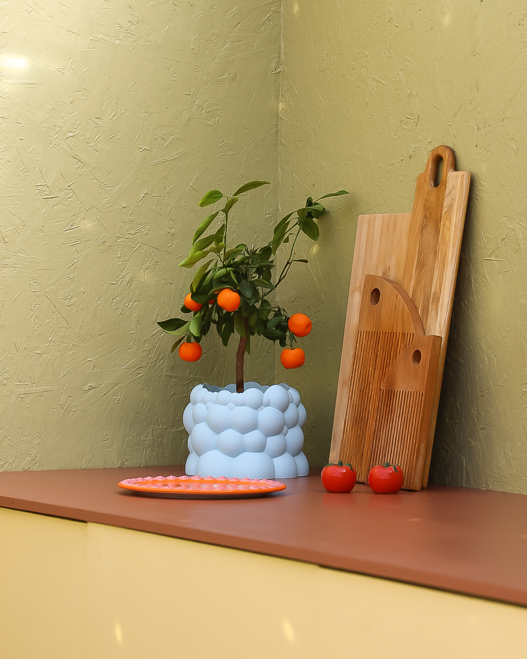 Detalj fra kjøkkenbenk med appelsinplante i lyseblå krukke og trefjøler, rød benkeplate i Jotun Amber Red, gult utekjøkken i Jotun Demigul og grønn vegg i Jotun Grønnland.