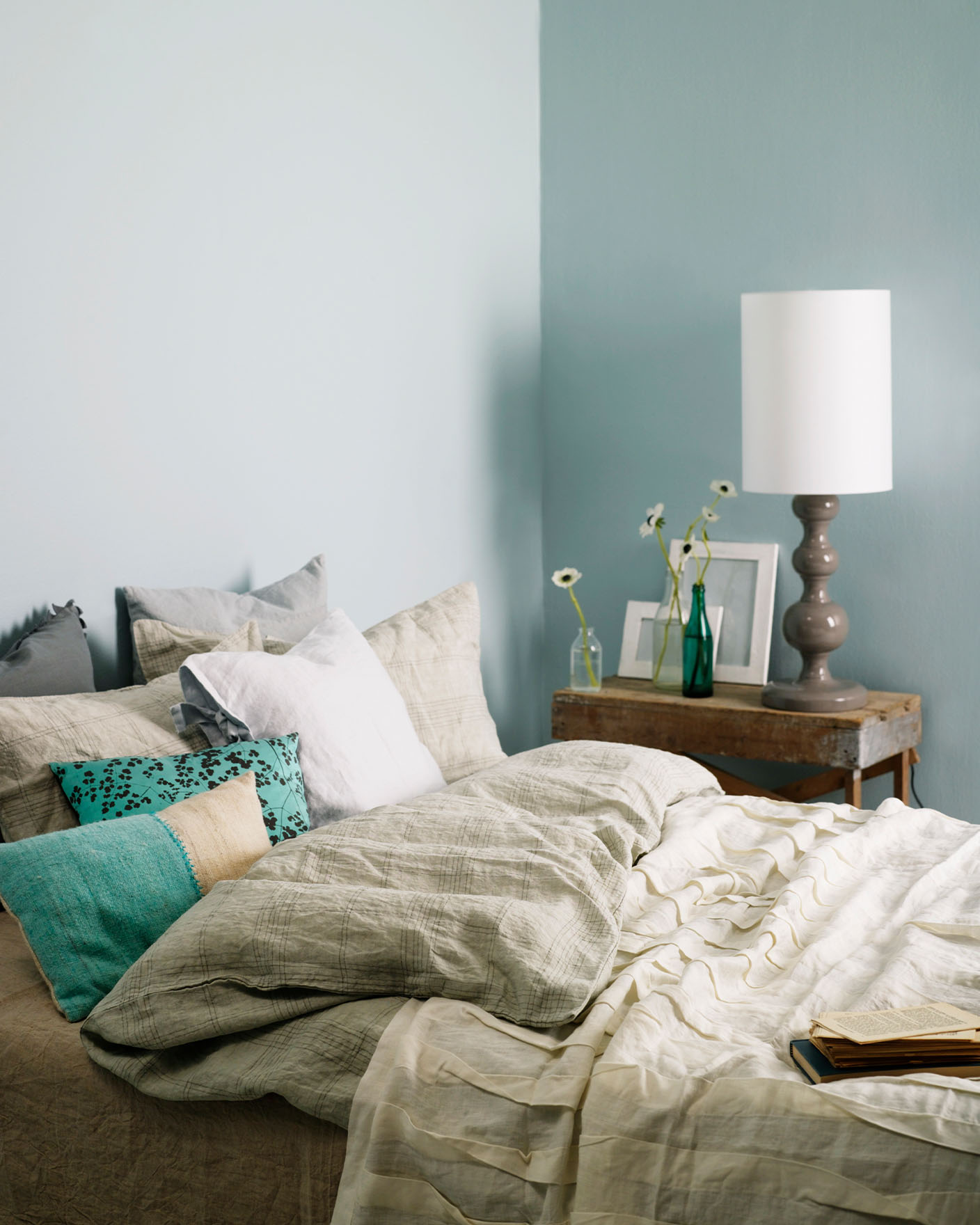 Inviting bedroom with layered neutral bedding, turquoise accent pillows, and a rustic wooden side table, set against muted, greenish blue wall painted with Jotun 5225 Lake View.