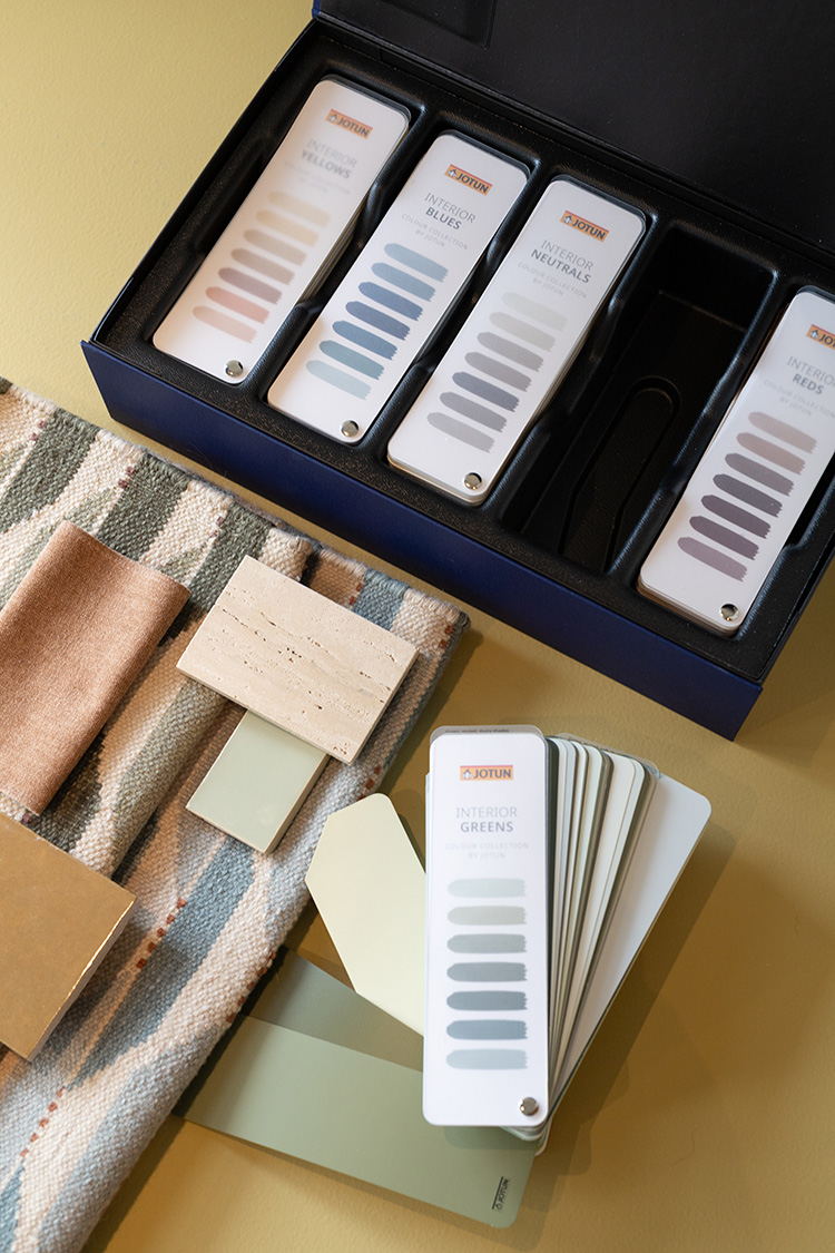 Fargeprøver og prøver som viser interiørdesignalternativer, inkludert malingsfarger fra Jotun i forskjellige nyanser som blått, grønt og nøytralt, vises sammen med teksturer av stoff og materialer på en beige overflate