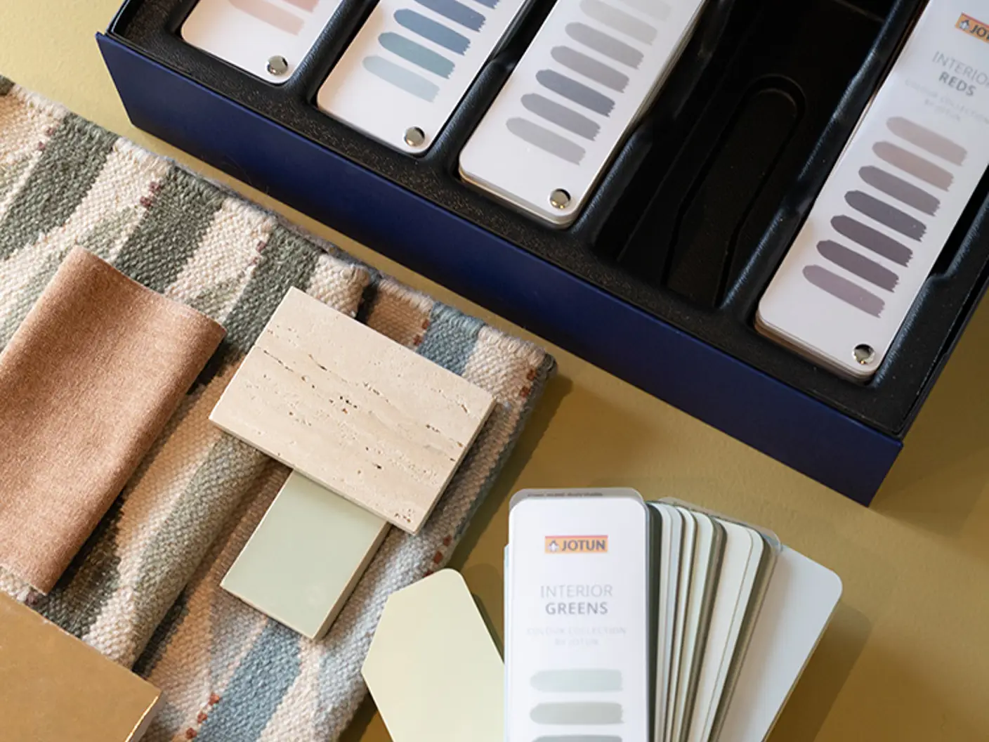 Fargeprøver og prøver som viser interiørdesignalternativer, inkludert malingsfarger fra Jotun i forskjellige nyanser som blått, grønt og nøytralt, vises sammen med teksturer av stoff og materialer på en beige overflate