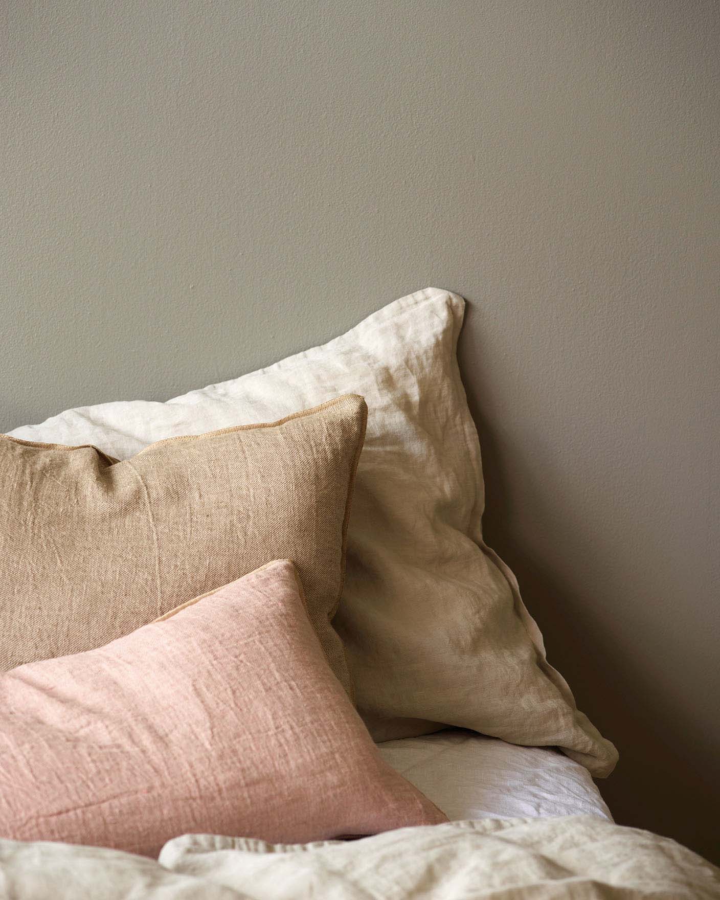 Close-up of a bed with linen pillows in soft beige, warm tan, and muted pink tones, set against a golden grey wall painted with Jotun 1877 Pebblestone.