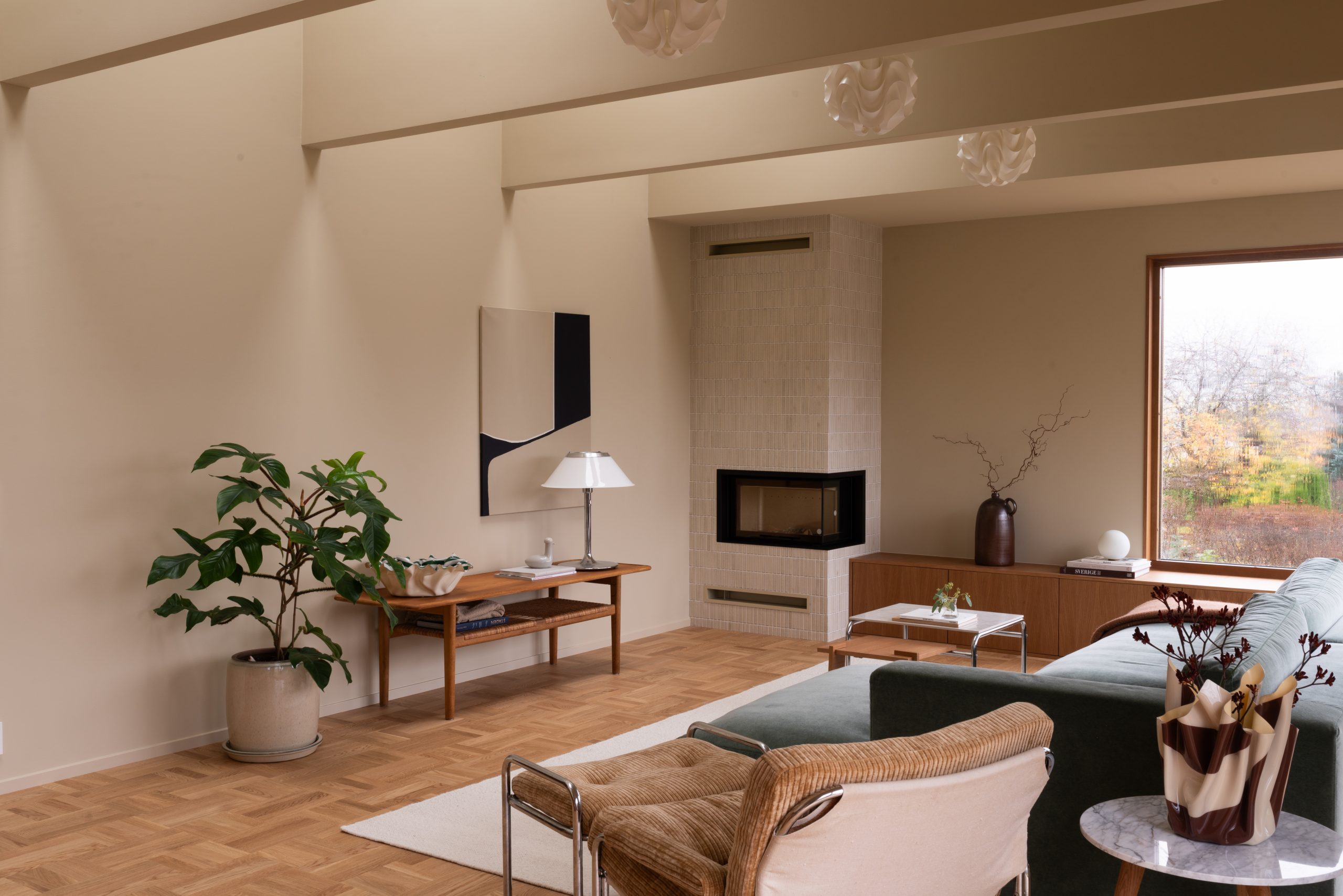 Lyst og moderne stue med parkettgulv, en sofa foran en stor vindu, to designstoler, og en peis. En plante står ved et sidebord med lampe og kunstverk. Minimalistisk skandinavisk interiør, vegger og tak malt i Jotun 11173 Humble Yellow