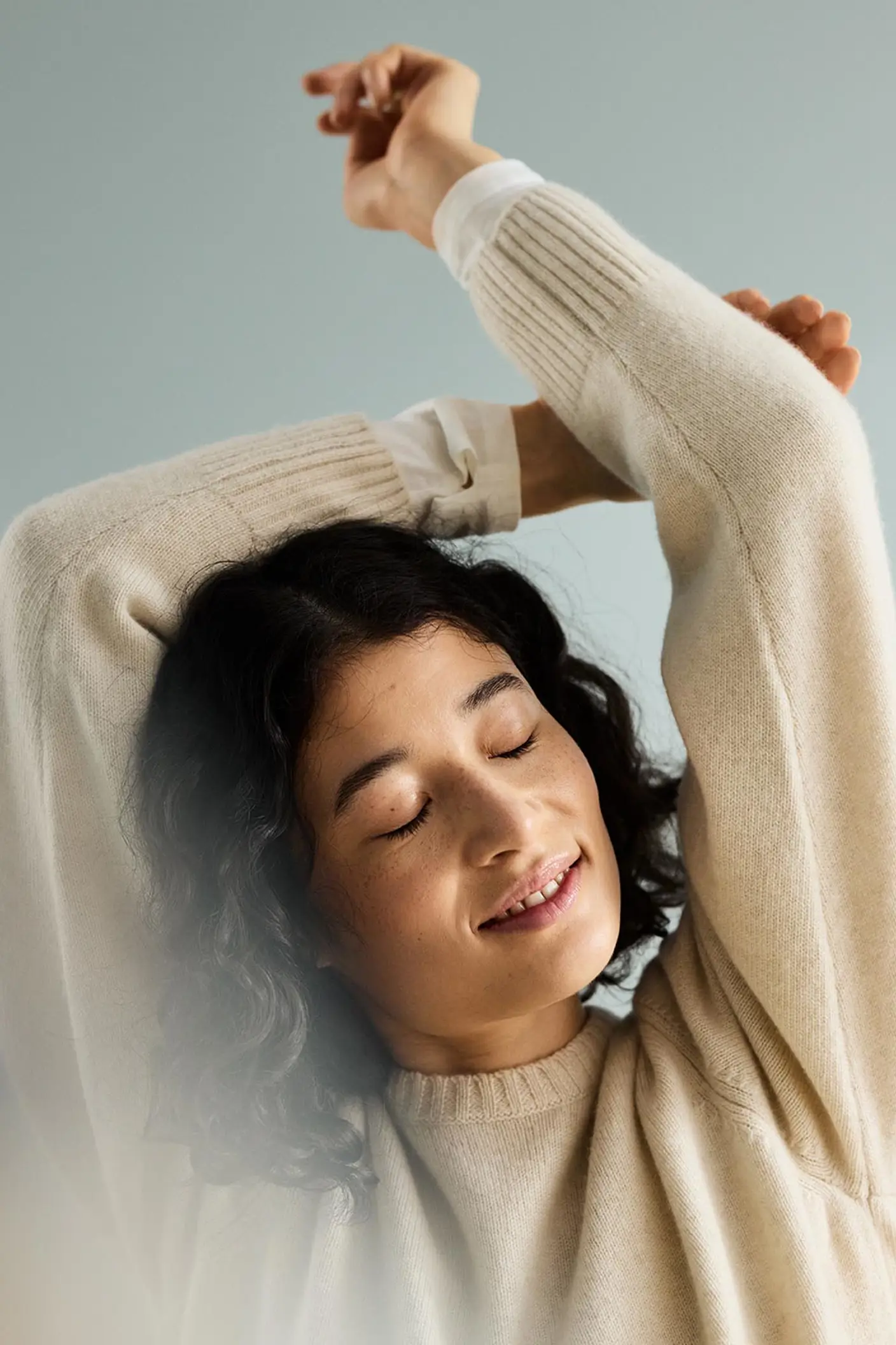 Woman with curly hair with arms raised above the head, in front of a muted blue background painted with the colour 6378 Iconic from Jotun.