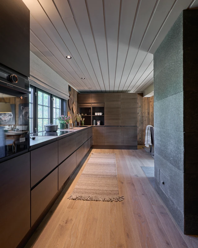 Moderne kjøkken med mørke treoverflater, stor benkplass, integrerte skap, og lyse tregulv. Vinduer gir naturlig lys, og innredningen har minimalistisk design. En løper dekker gulvet