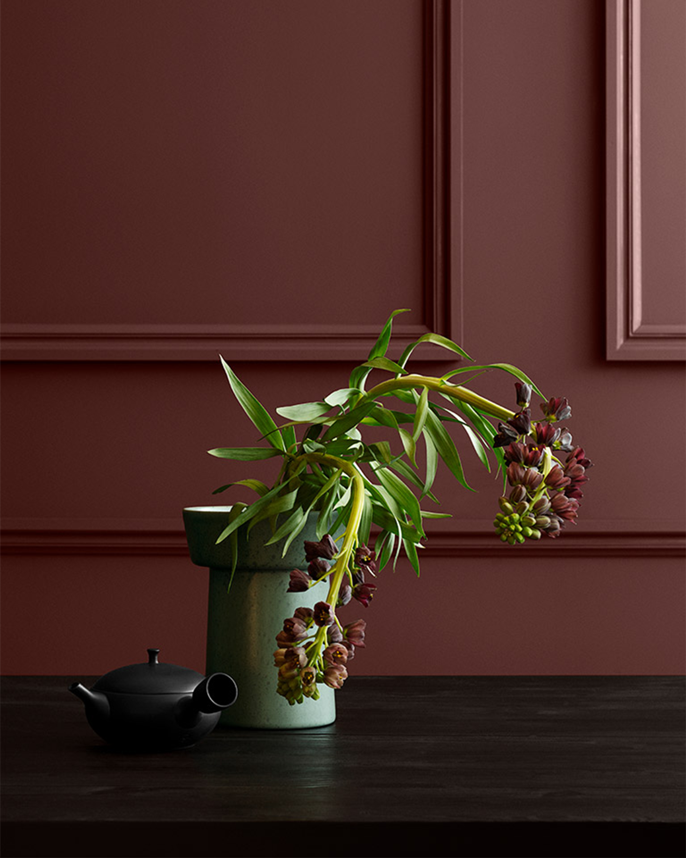 Grønn blomstergren i lysegrønn vase på mørkt bord, med svart tekanne mot burgunder vegg malt i Jotun Red Maple.