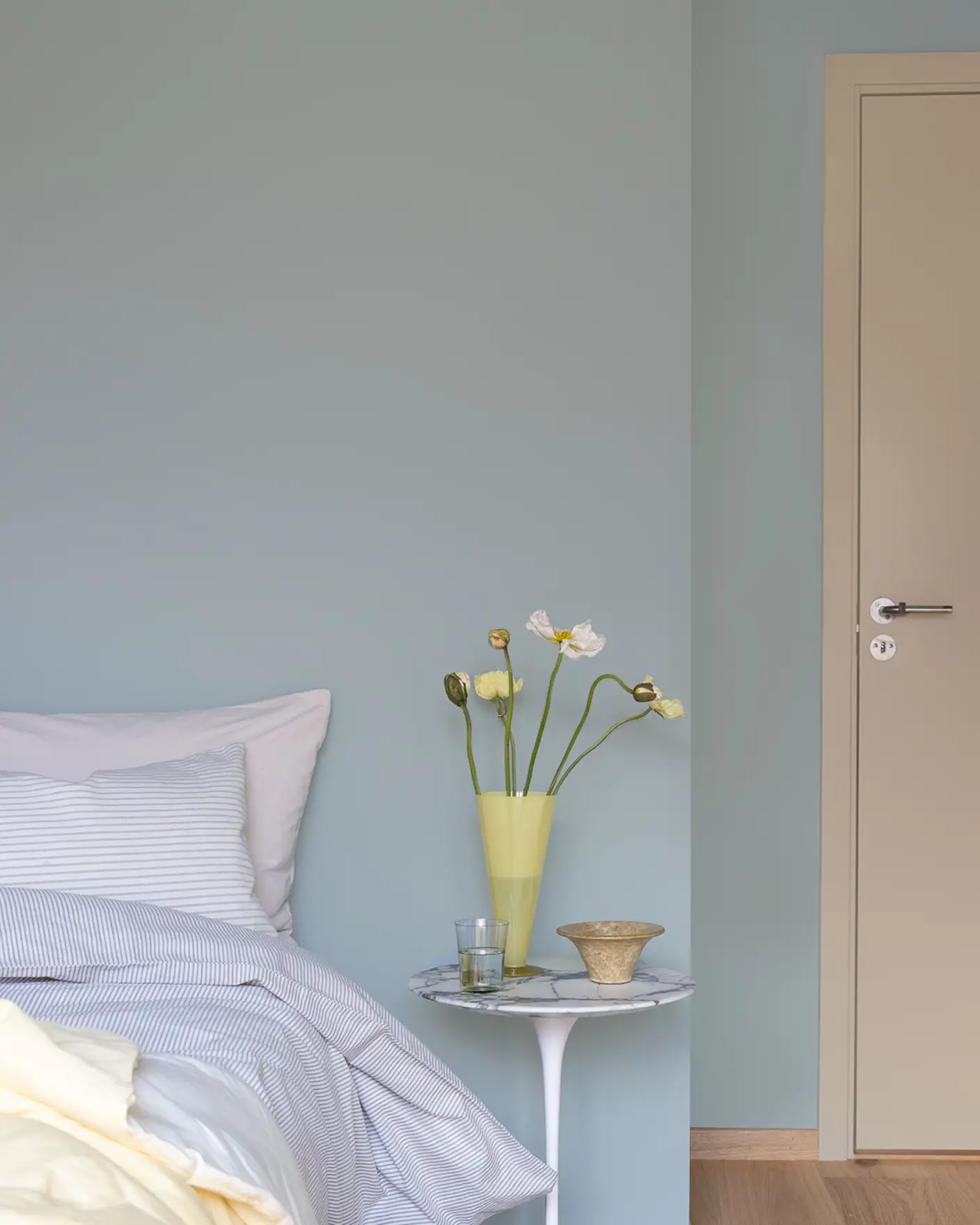 Bedroom with fresh linen and yellow flowers in nightstand, walls painted in buegreen JOTUN 6378 Iconic and door with trimmings in dark beige JOTUN 12300 Hazelnut Beige.