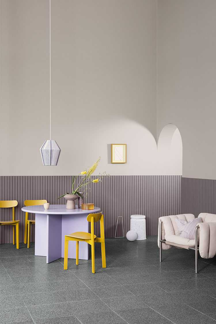 Moderne interiørdesign med et lilla og gult fargevalg som inneholder et rundt lavendelfarget bord, gule stoler, pendellampe og en koselig hvit lenestol mot et dekorativt veggpanel malt lilla i fargen Jotun 3377 Slate Lavender
