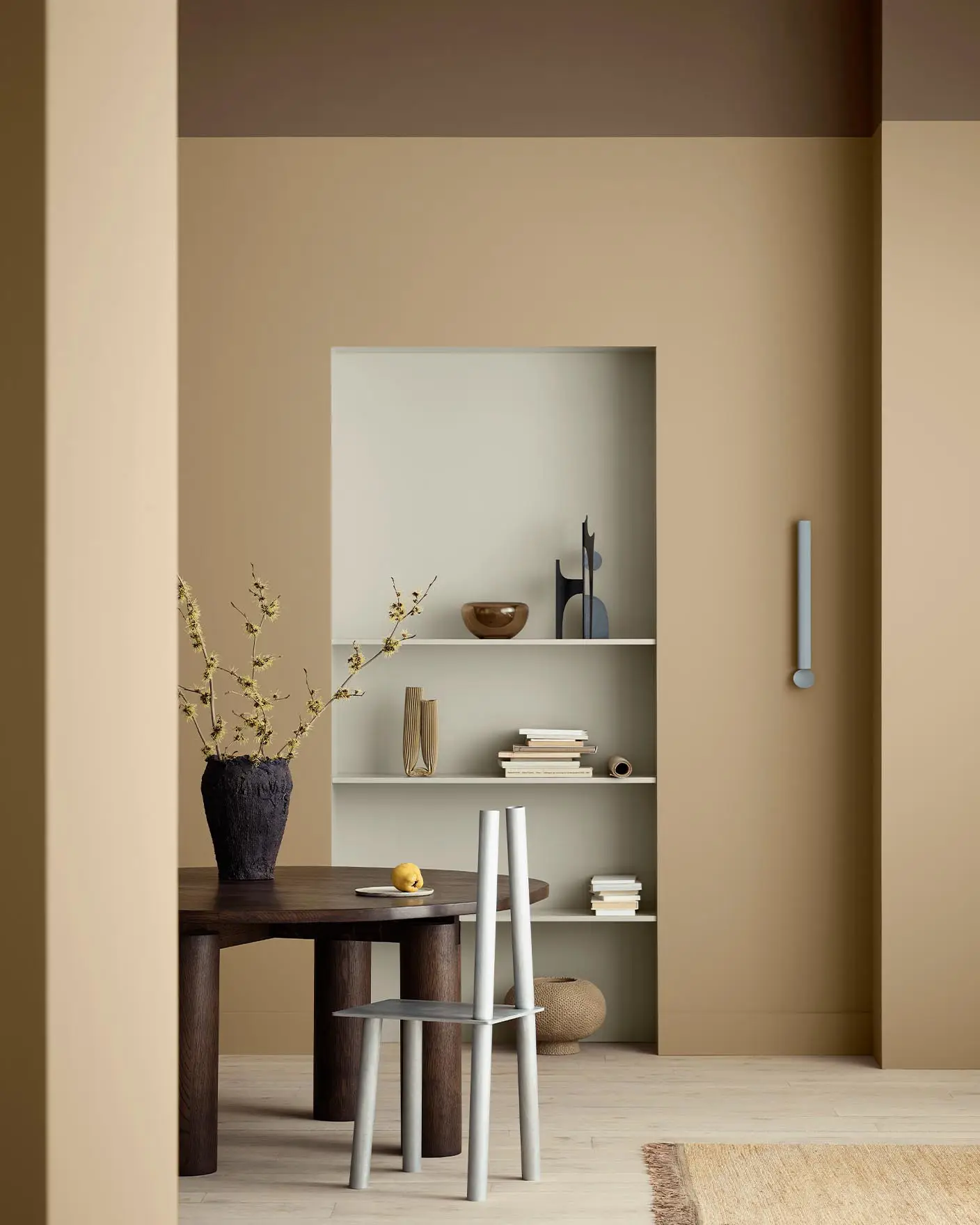 A warm, minimal dining space with golden brown walls painted with Jotun 1632 Rock Sugar, and built-in shelving, painted with Jotun 12075 Soothing Beige, styled with books and ceramics.