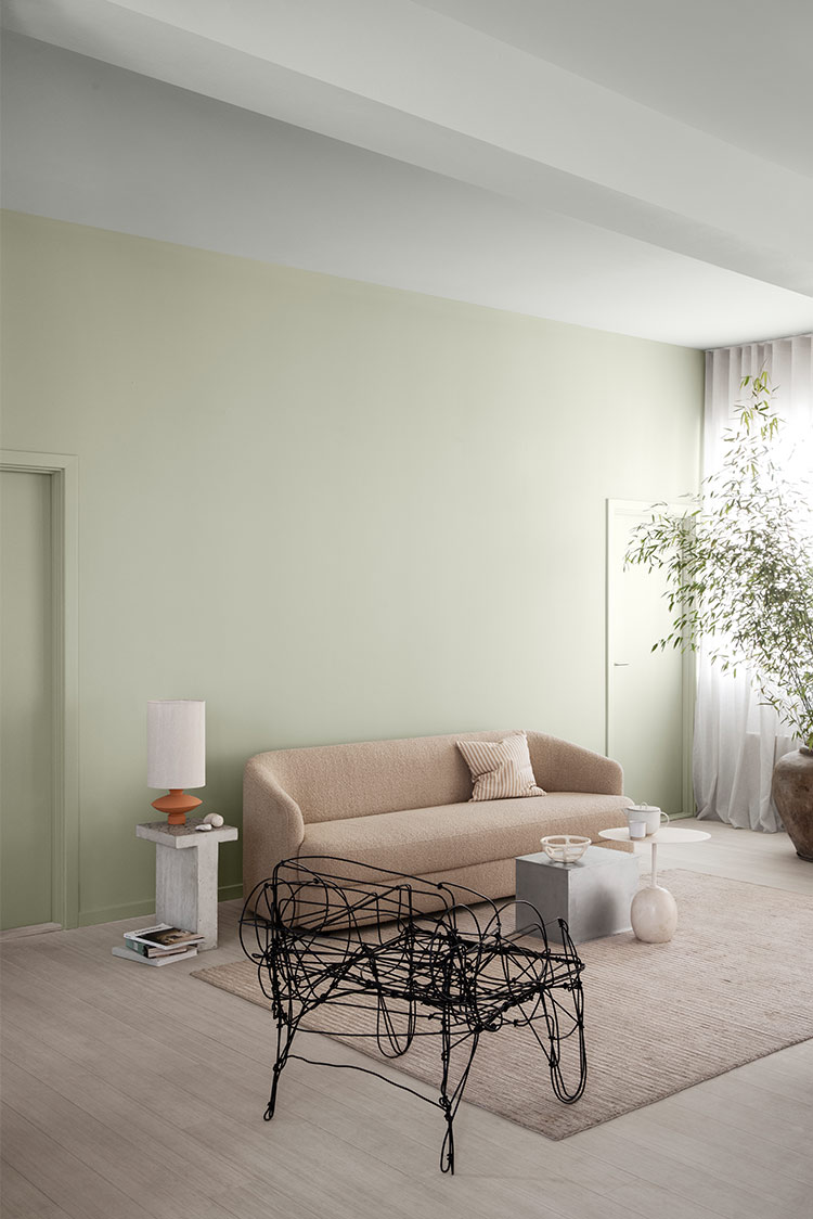 Moderne stue med en beige sofa, minimalistisk sidebord, abstrakt trådstol og en potteplante, med et nøytralt fargevalg og naturlig lys på lysegrønne vegger malt i Jotun 8281 Pale Linden