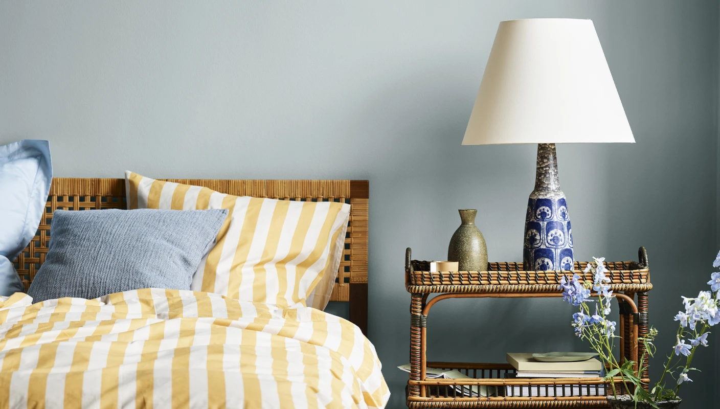 Cozy bedroom with yellow and white striped bedding and a wicker nightstand with a blue wall painted with 6236 Darjeleling from Jotun.