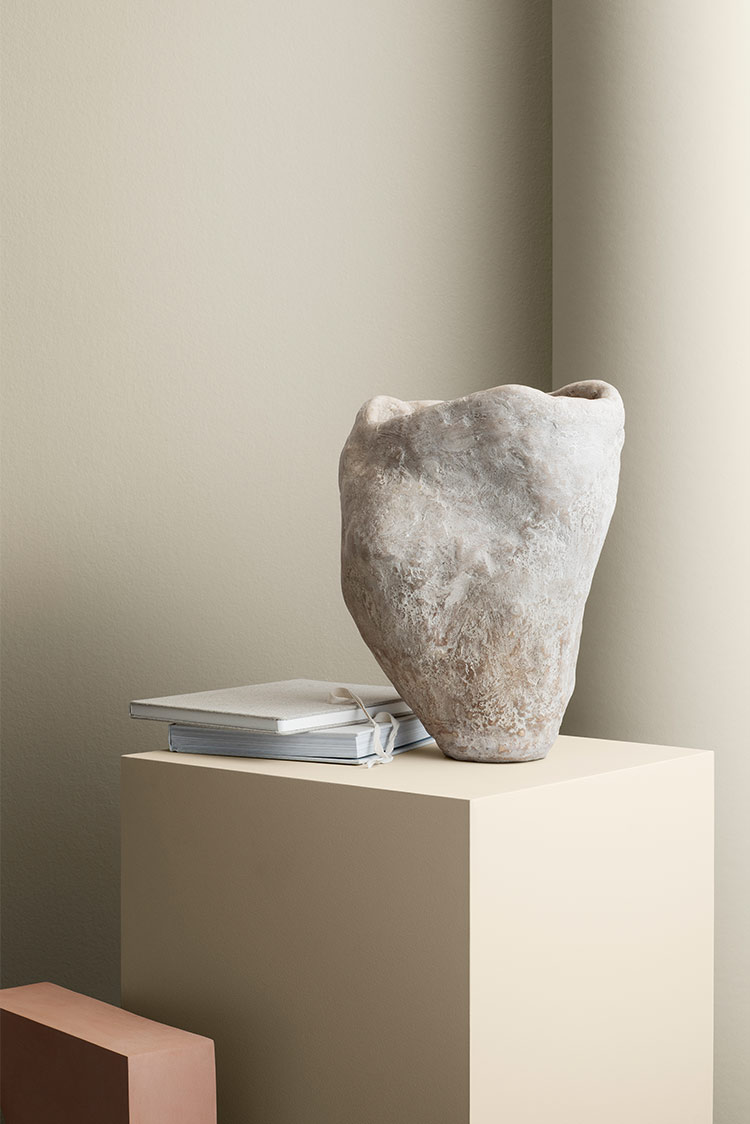 Rustikk steinvase og lukkede notatbøker på beige sokkel i minimalistisk setting med beige vegg i bakgrunnen malt i Jotun 12182 Gentle Whisper