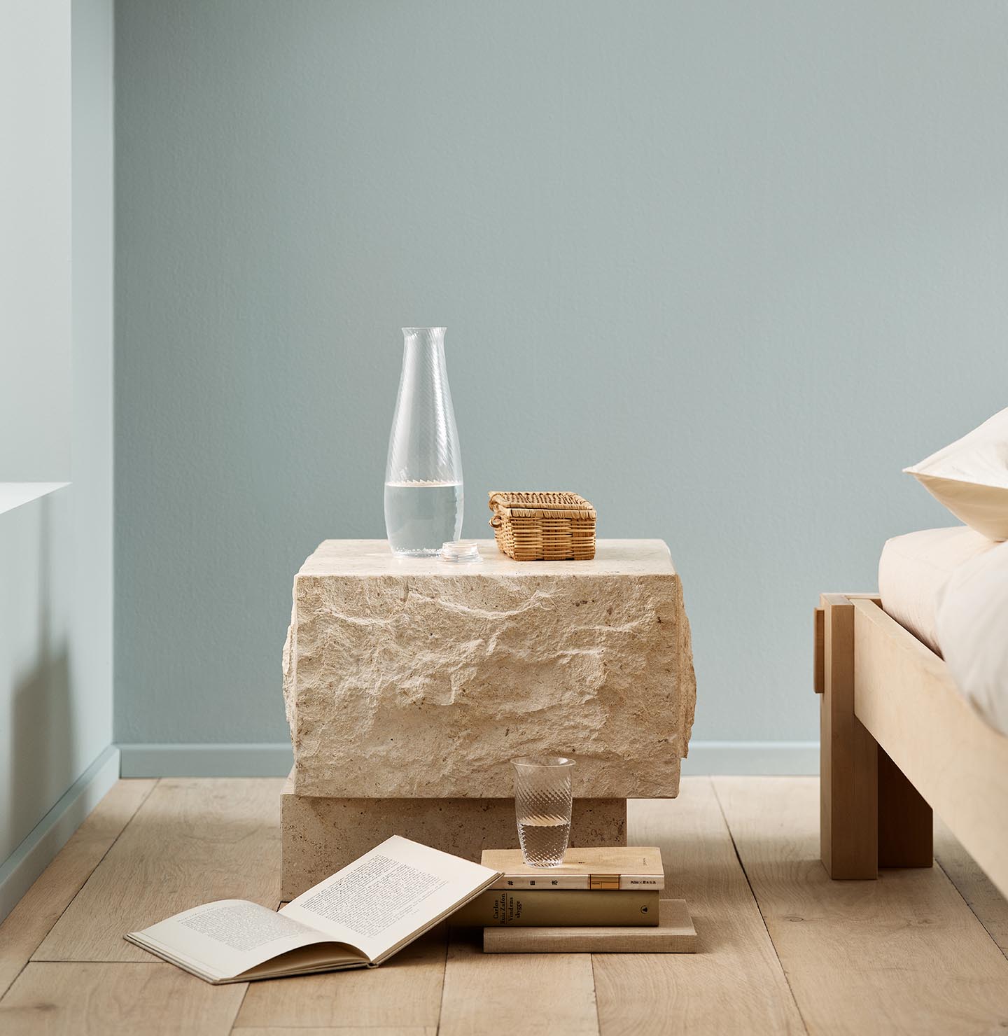Minimalist bedroom with a stone bedside table and books on a wooden floor against a muted blue wall painted with 6378 Iconic from Jotun.