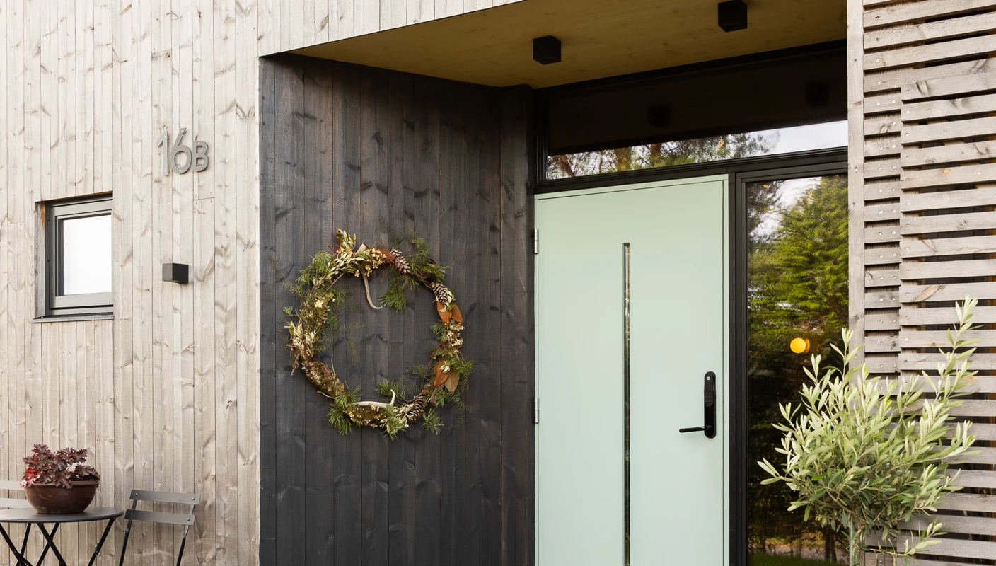 Inngangsparti til moderne funkishus i malmfuru, vegg rundt lysegrønn dør oljet med TREBITT WoodCare i gråsorte Muted Charcoal og tak over dør i gylne Fair Wood.