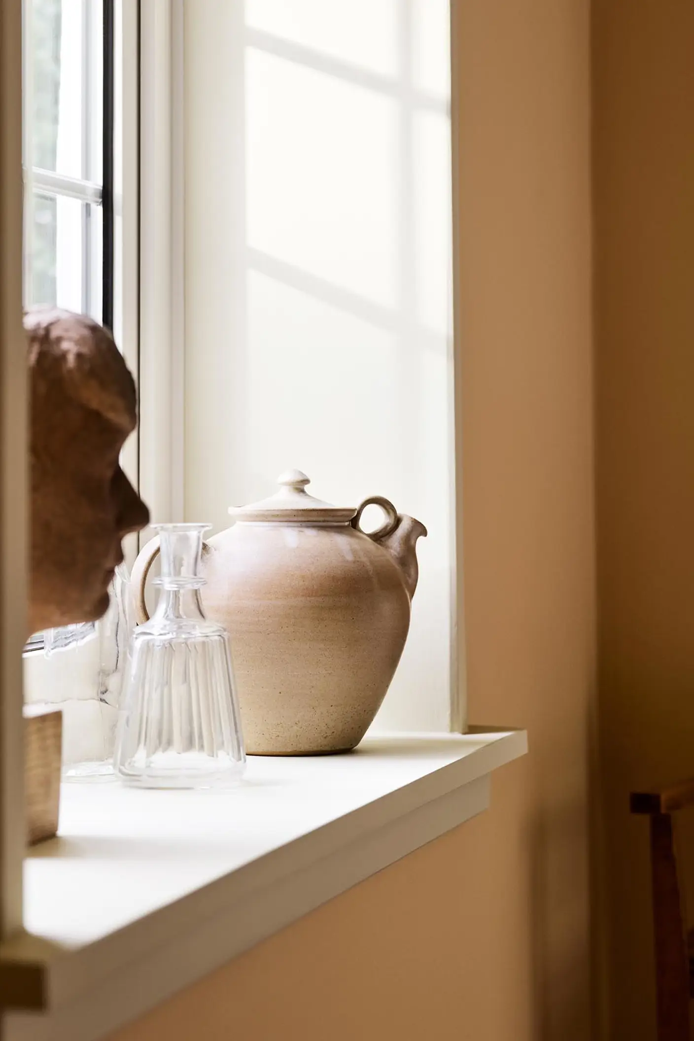 Ceramic teapot and glass pitcher with sunlight against a ochre yellow wall painted in the Jotun colour 11202 Mild Ochre.