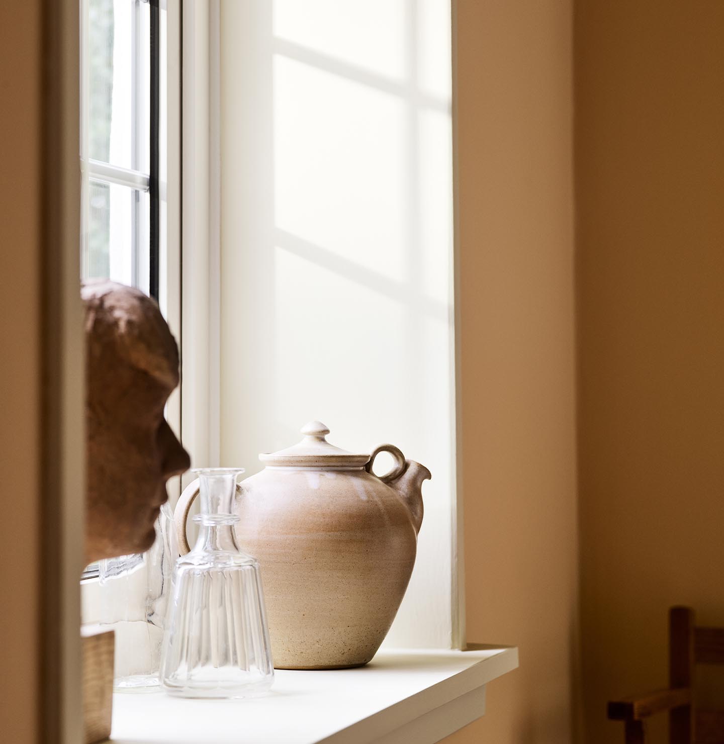 Ceramic teapot and glass pitcher with sunlight against a ochre yellow wall painted in the Jotun colour 11202 Mild Ochre.