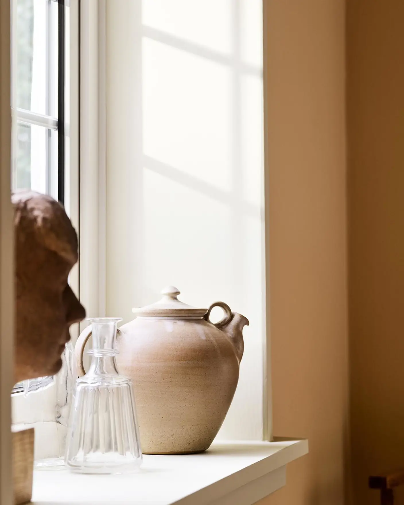Ceramic teapot and glass pitcher with sunlight against a ochre yellow wall painted in the Jotun colour 11202 Mild Ochre.