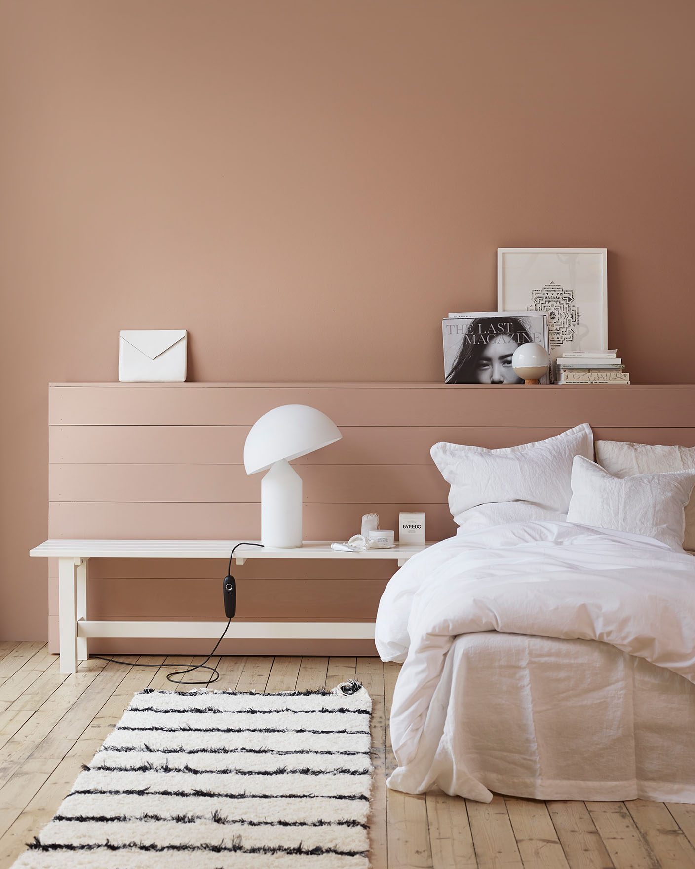 Bedroom with a white linen bed, striped rug, a modern white lamp on a bench, set against muted, golden pink wall painted with Jotun 2024 Senses.