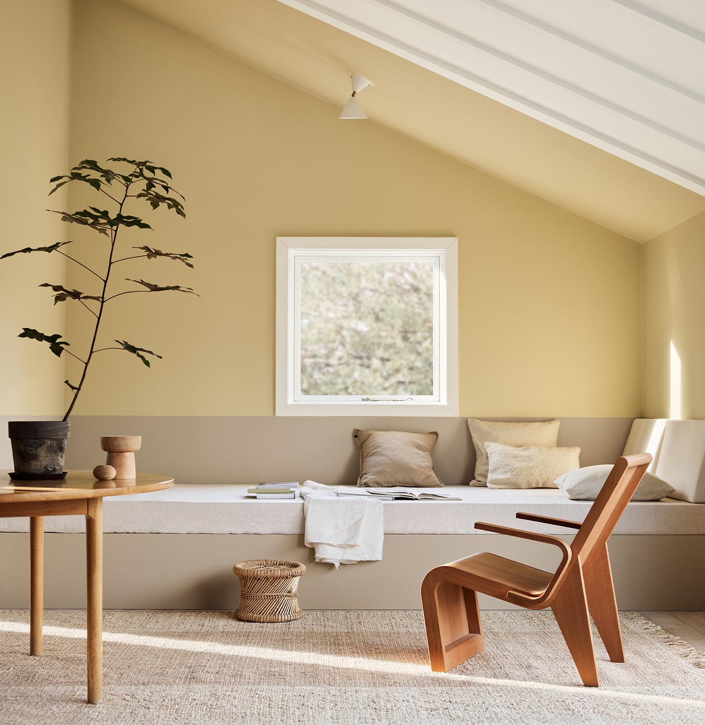 A daybed, wodden chair and a potted plant against muted, greenish yellow walls painted in the colour 1965 Ginger Tea from Jotun. Daybed painted in the colour 12300 Hazelnut Beige. 
