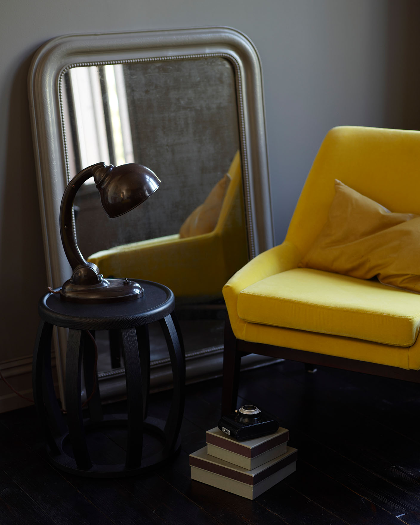 Mustard yellow chair, vintage mirror, dark wood side table with an industrial lamp and stacked boxes, all set against a brownish grey wall painted with Jotun 2732 Deep Mocca.