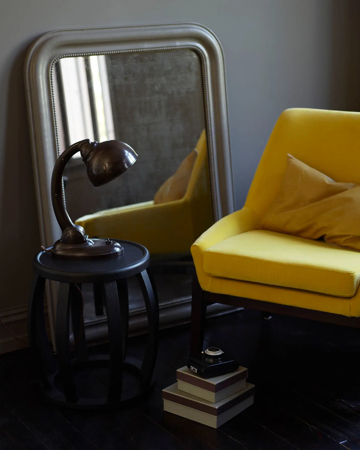 Mustard yellow chair, vintage mirror, dark wood side table with an industrial lamp and stacked boxes, all set against a brownish grey wall painted with Jotun 2732 Deep Mocca.