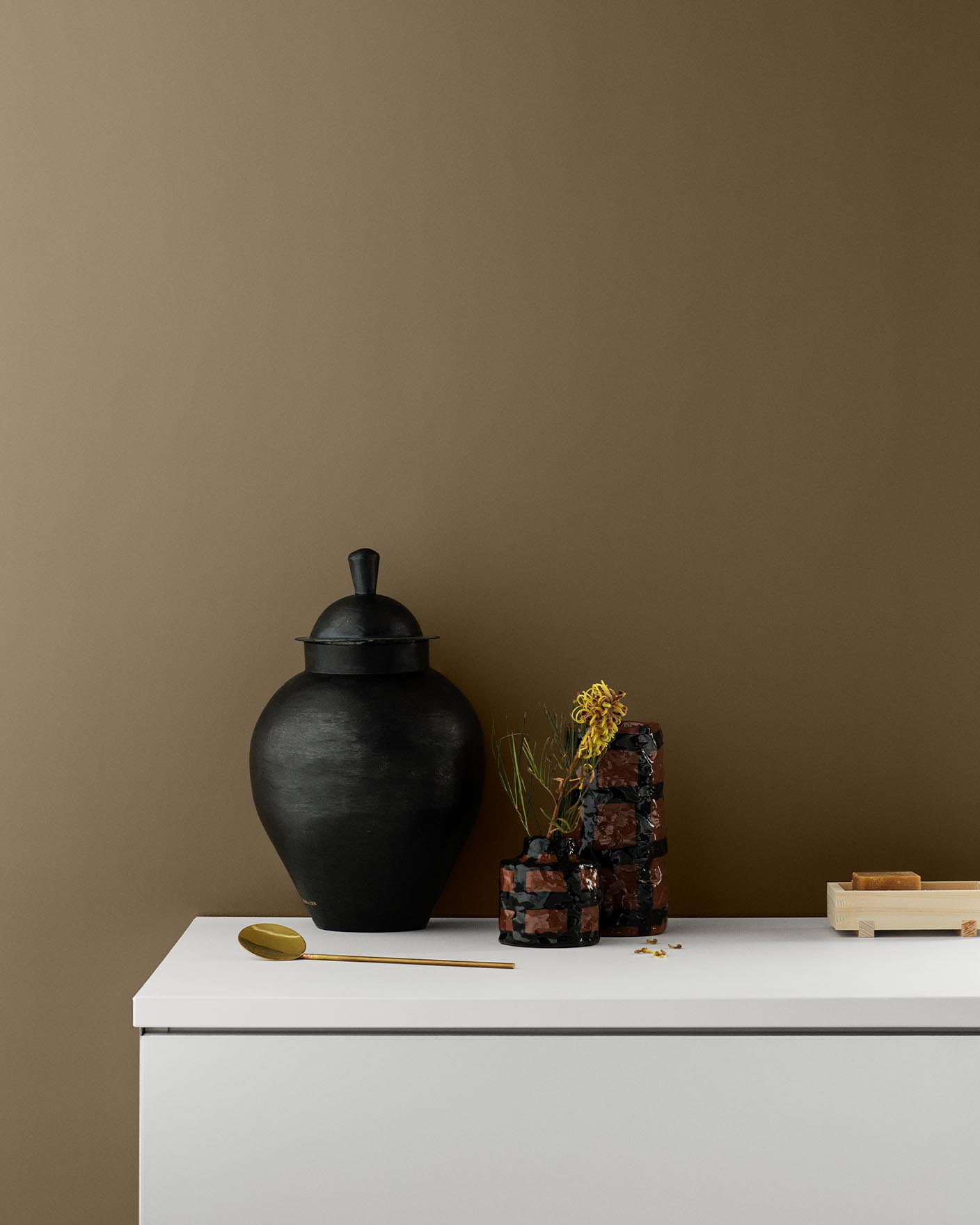Minimalistisk innredning med svart vase og planter på en hvit overflate mot en gyldenbrun vegg malt med fargen 10693 Golden Bronze fra Jotun.