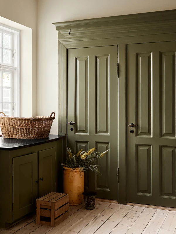 Lyst interiør med olivengrønne dører malt i Jotun 8597 Seaweed Green, matchende skap, kurv på benk, og gul vase med dekorative planter, på lyst tregulv