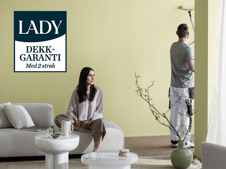 En kvinne sitter i en sofa i en stue og en mann maler veggen i en lys grønnaktig gul farge.