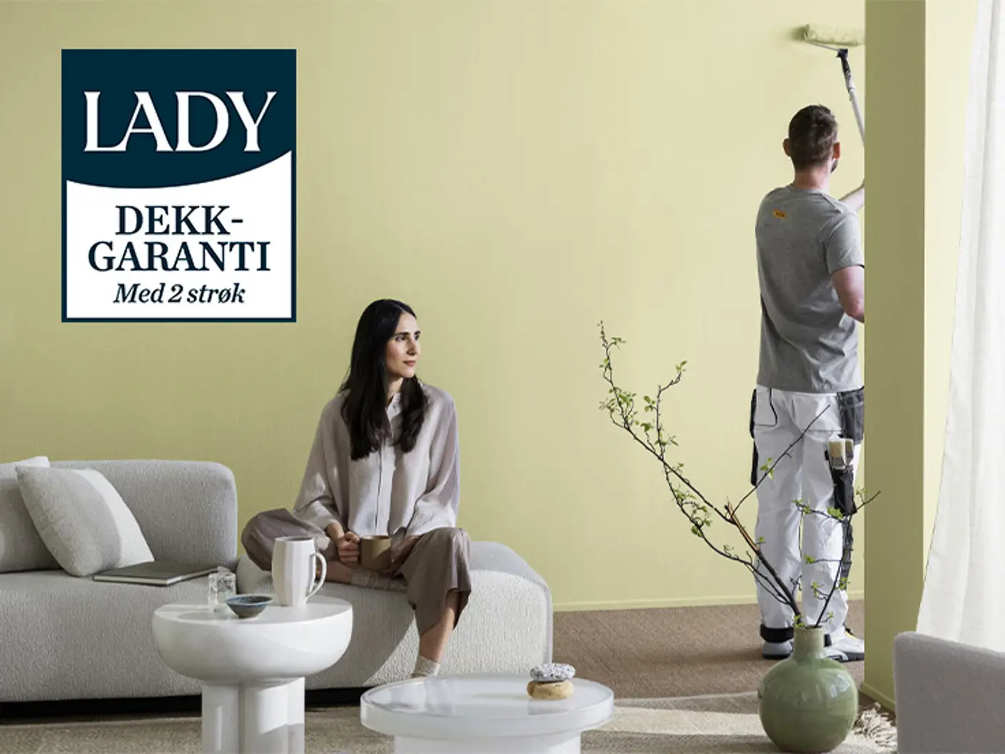 En kvinne sitter i en sofa i en stue og en mann maler veggen i en lys grønnaktig gul farge.